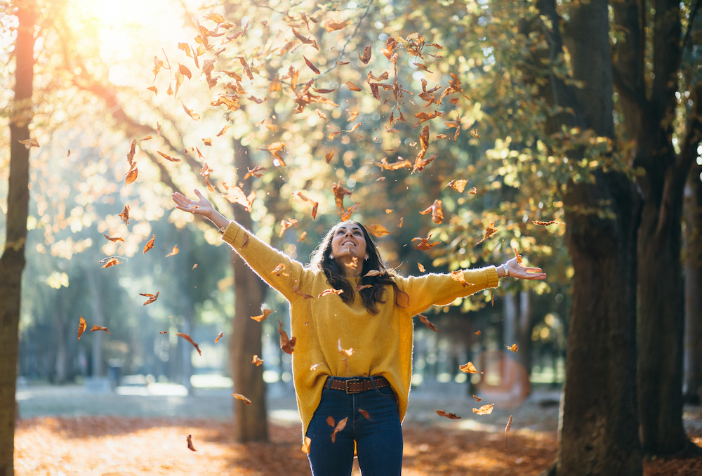 Casual joyful woman having fun throwing leaves in autumn at city park.