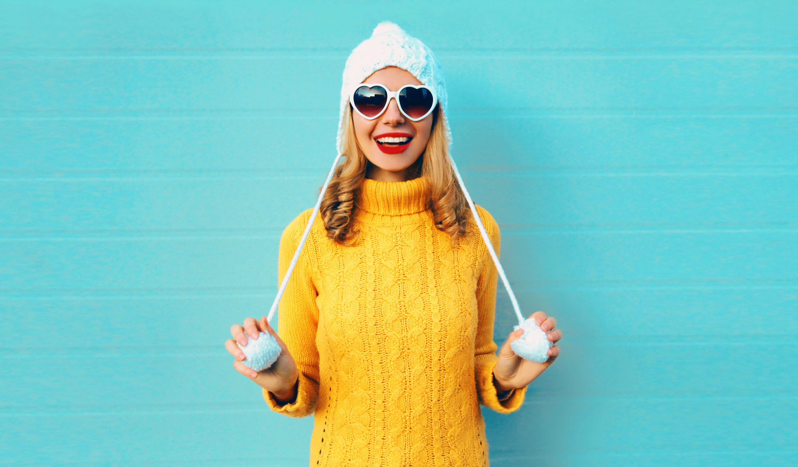 Winter portrait happy smiling young woman having fun wearing yellow knitted sweater and white hat with pom pom, heart shaped sunglasses on blue wall background