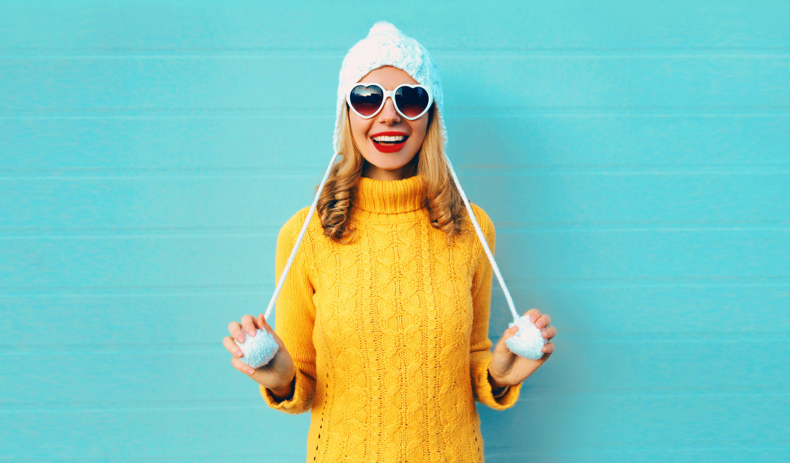 Winter portrait happy smiling young woman having fun wearing yellow knitted sweater and white hat with pom pom, heart shaped sunglasses on blue wall background Winter portrait happy smiling young woman having fun wearing yellow knitted sweater and white hat with pom pom, heart shaped sunglasses on blue wall background