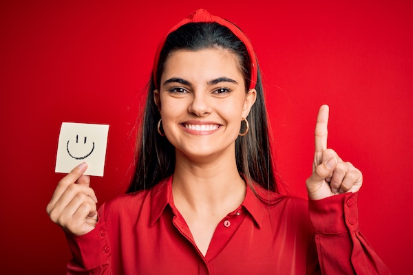 Young beautiful brunette woman holding reminder paper with smile emoji message surprised with an idea or question pointing finger with happy face, number one